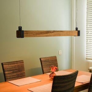 Rechthoekige hanglamp van massief hout met strak afgewerkte zwarte metalen eindkappen en geïntegreerde LED-lichtstrip aan de onderzijde. De lamp verspreidt warm wit licht en hangt aan staalkabels, ideaal als moderne verlichting boven een eettafel in een eigentijds of industrieel interieur.