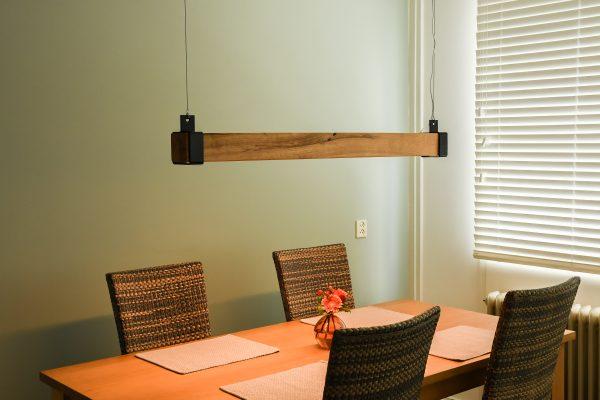 Rechthoekige hanglamp van massief hout met strak afgewerkte zwarte metalen eindkappen en geïntegreerde LED-lichtstrip aan de onderzijde. De lamp verspreidt warm wit licht en hangt aan staalkabels, ideaal als moderne verlichting boven een eettafel in een eigentijds of industrieel interieur.